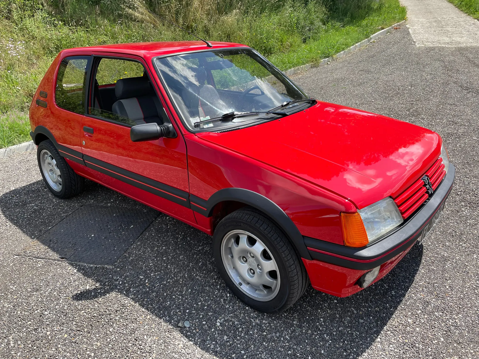 Peugeot 205 rouge vif à bandeau noir, carrosserie brillante en plein soleil